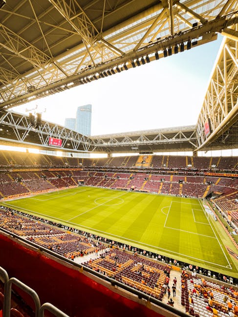 Inside a large stadium with a view of a well-maintained grass football pitch, surrounded by tiered seating areas with red and yellow seats, some of which display the word 'BRENT'. Overhead, the stadiu
